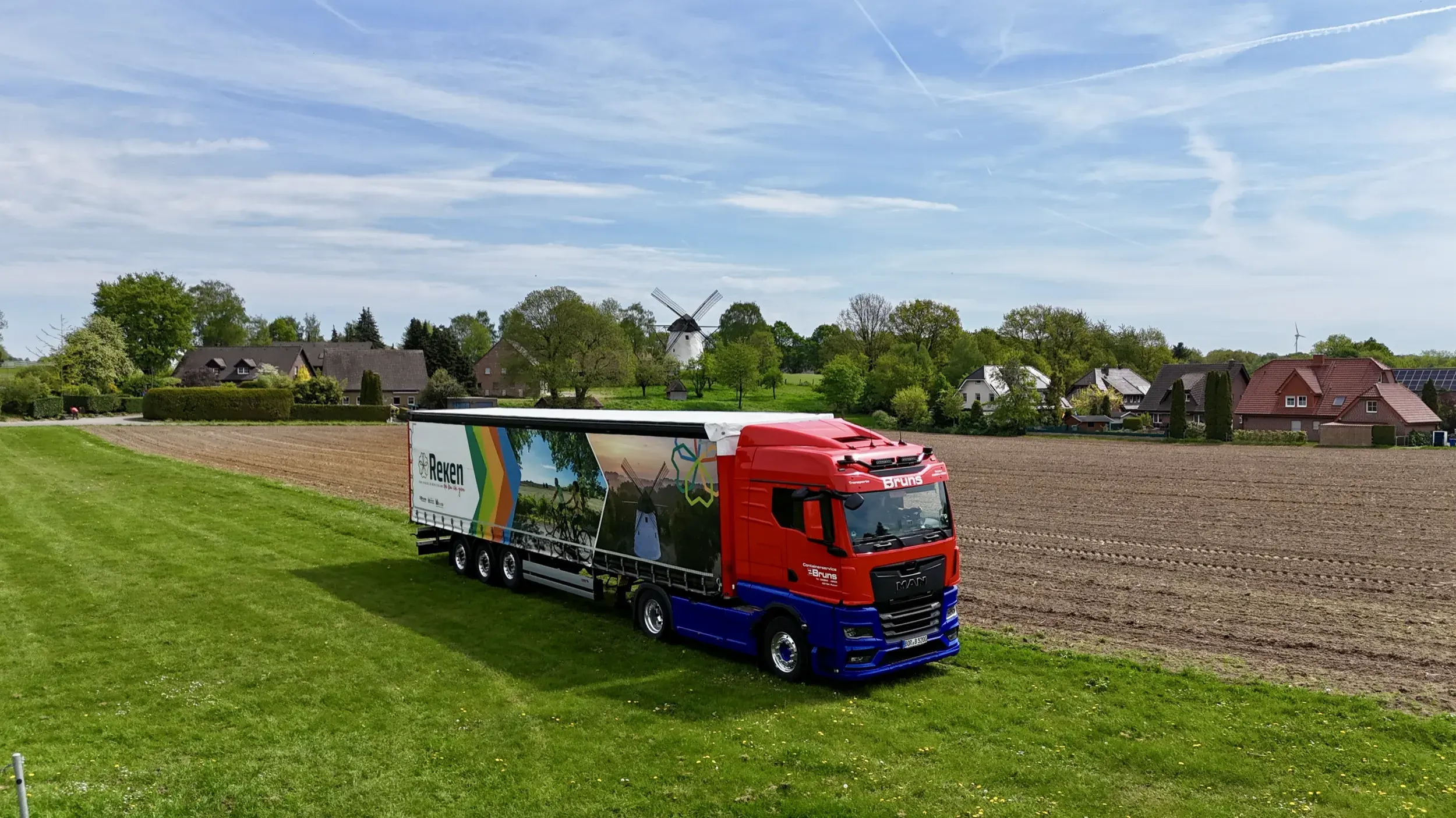 Luftaufnahme: Sattelzug mit bedrucktem Planenauflieger auf einem Feld, im Hintergrund eine Windmühle